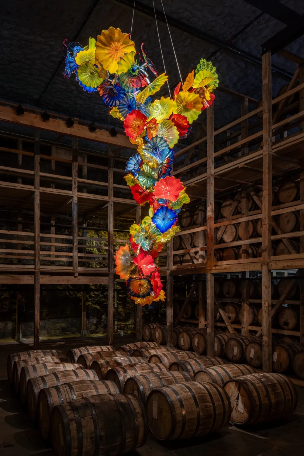 A vibrant, colorful glass sculpture hangs from the ceiling, surrounded by wooden barrels in a dimly lit cellar.