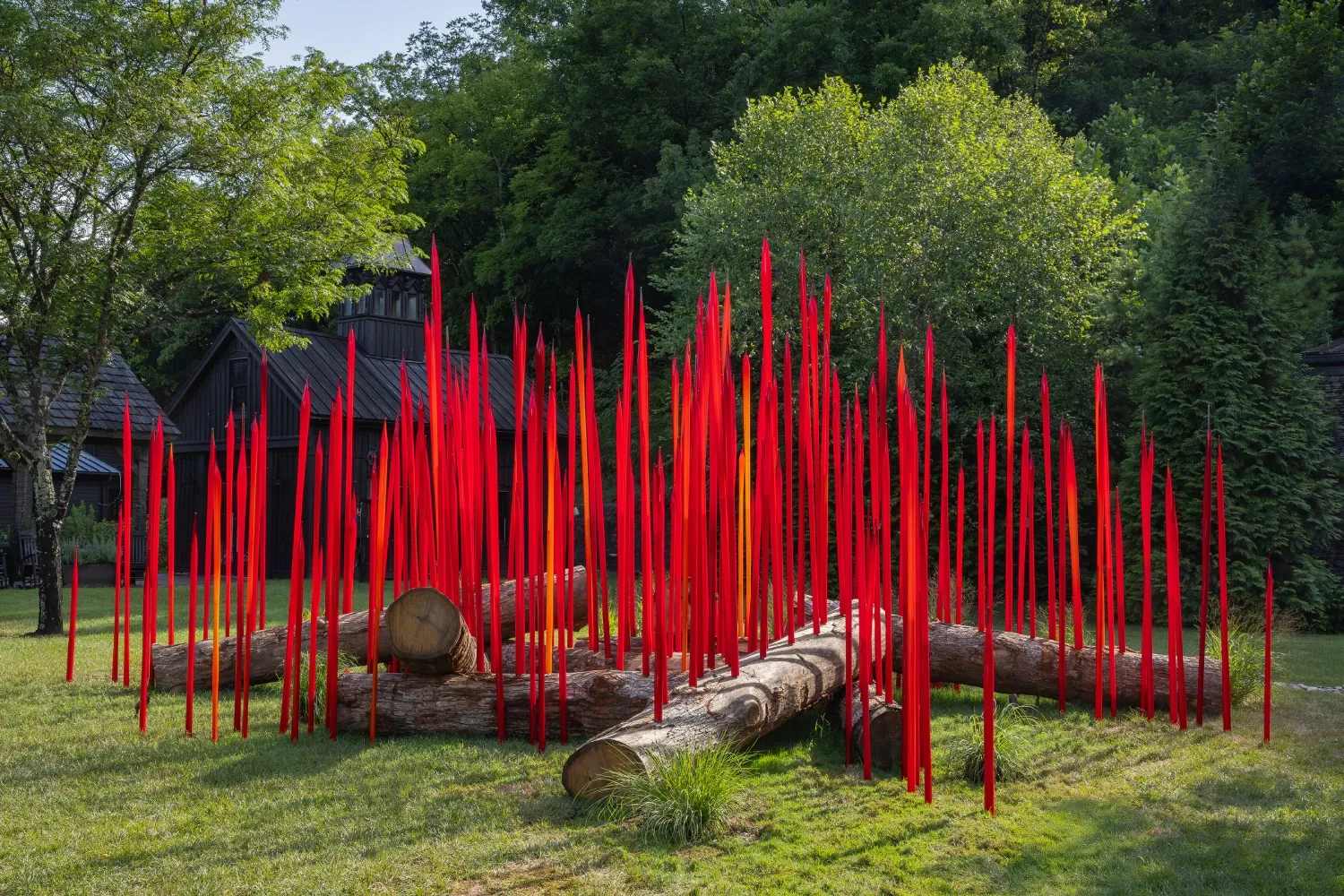 A colorful outdoor sculpture featuring tall red glass spikes arranged around wooden logs, surrounded by lush greenery.